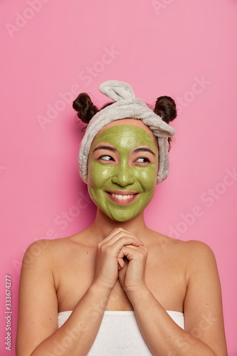 Fototapeta Naklejka Na Ścianę i Meble -  Pleased Asian woman undergoes treatment with facial mask, smiles toothily, keeps hands together, wrapped in towel after taking shower, wears headband, poses indoor. People, rejuvenation concept