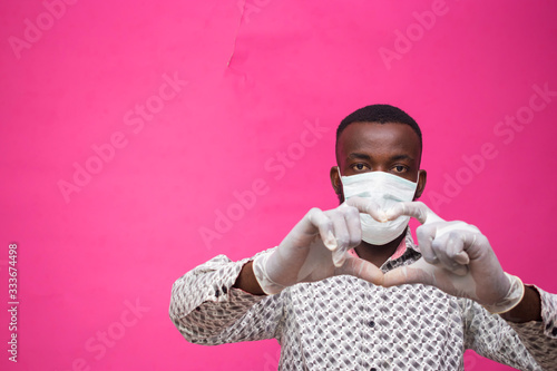 a young african doctor isolated over pink background wearing face mask to prevent, preventing, prevented himself from the outbreak in the society and did a love sign.