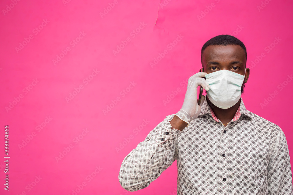 a young african doctor isolated over pink background wearing face mask to prevent, prevented, preventing himself from the outbreak in the society, making call with his smartphone.