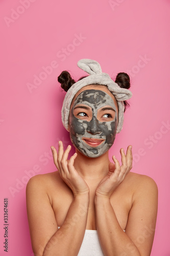 Portrait of good looking Asian woman applies clay mask on face, stands wrapped in towel, looks aside, has smooth perfect skin, takes treatment at spa salon, isolated on pink studio wall. Wellness