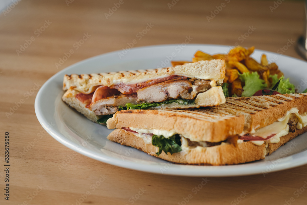 Close-up of chicken and bacon club sandwich with fries and vegetables on a white plate on a wooden background.