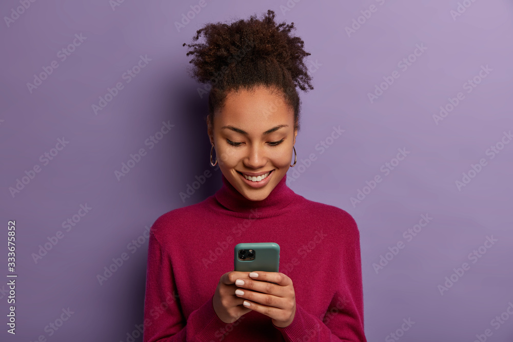 Happy beautiful Afro American woman uses social media messanger on modern mobile phone, chats with friends, going to have video call, dressed in casual wear, makes shopping in internet store - obrazy, fototapety, plakaty 