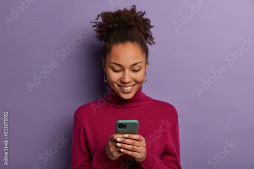Happy beautiful Afro American woman uses social media messanger on modern mobile phone, chats with friends, going to have video call, dressed in casual wear, makes shopping in internet store