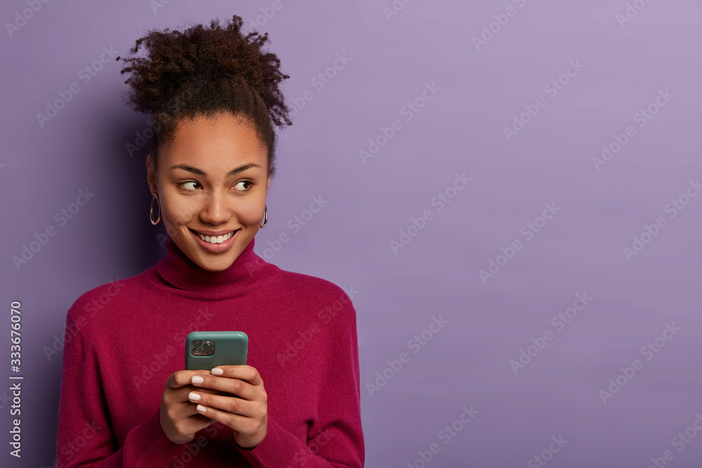 Smiling pleasant looking woman receives message from dating app, holds mobile phone in hands, looks happily aside, edits photo to post online, has curly hair, isolated on violet wall, copy space Smiling pleasant looking woman receives message from dating app, holds mobile phone in hands, looks happily aside, edits photo to post online, has curly hair, isolated on violet wall, copy space