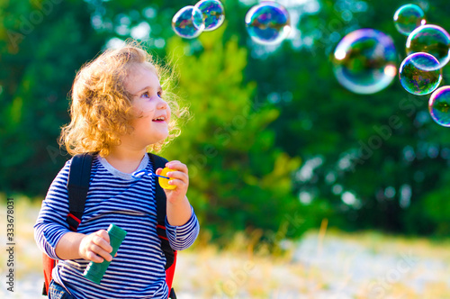 horizontal portrait of a cheerful curly-haired girl with a red backpack and soap bubbles on a background of nature