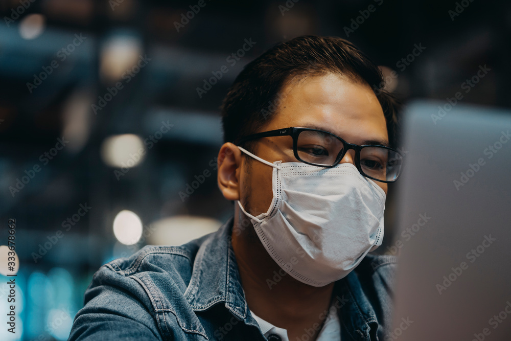 Asian young man indoors in cafe. Lifestyle concept photo with copy space. Picture with handsome guy who using protective face mask. Portrait with gray laptop