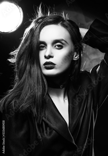 Beautiful slim girl model with red lips, wearing a black blazer, touches her hair with her hand on a dark background, posing next to a light lamp. Advertising, monochrome, black and white design. © Алексей Торбеев
