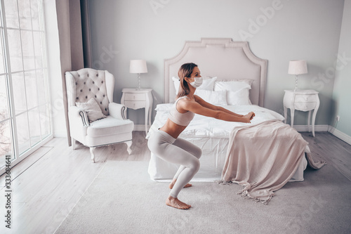 Home fitness. Young fit slim woman in protection face mask doing morning work-out yoga exercises in bedroom during self isolation quarantine. COVID-19 concept to promote stay safe home save lives
