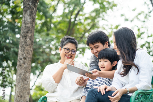Asian happy family play in park, read electronic book from tablet together. Boy sit in on bench between grandmother and mother. Father hug him from back, point interest thing. Family lifestyle concept