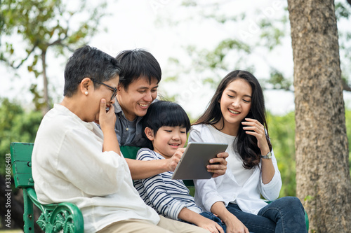 Asian happy family play in park, read electronic book from tablet together. Boy sit in on bench between grandmother and mother. Father hug him from back, point interest thing. Family lifestyle concept © Kawee