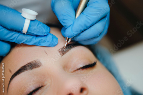 Permanent make-up for eyebrows of beautiful brunette woman in beauty salon. Closeup beautician in gloves doing microblading for eyebrow. photo