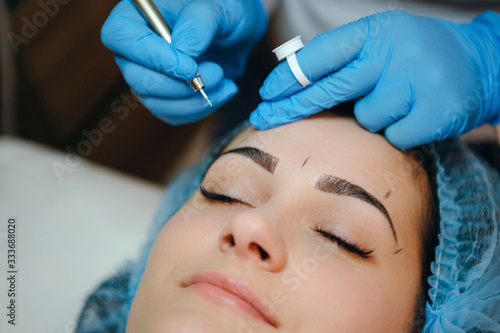 Permanent make-up for eyebrows of beautiful brunette woman in beauty salon. Closeup beautician in gloves doing microblading for eyebrow.