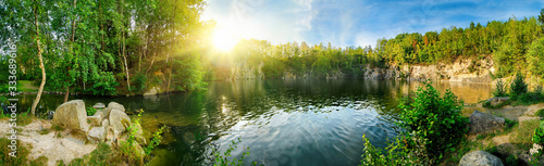 Fototapeta Naklejka Na Ścianę i Meble -  Panoramic landscape shot of idyllic lake surrounded by trees and cliffs, with the sun glowing on the horizon