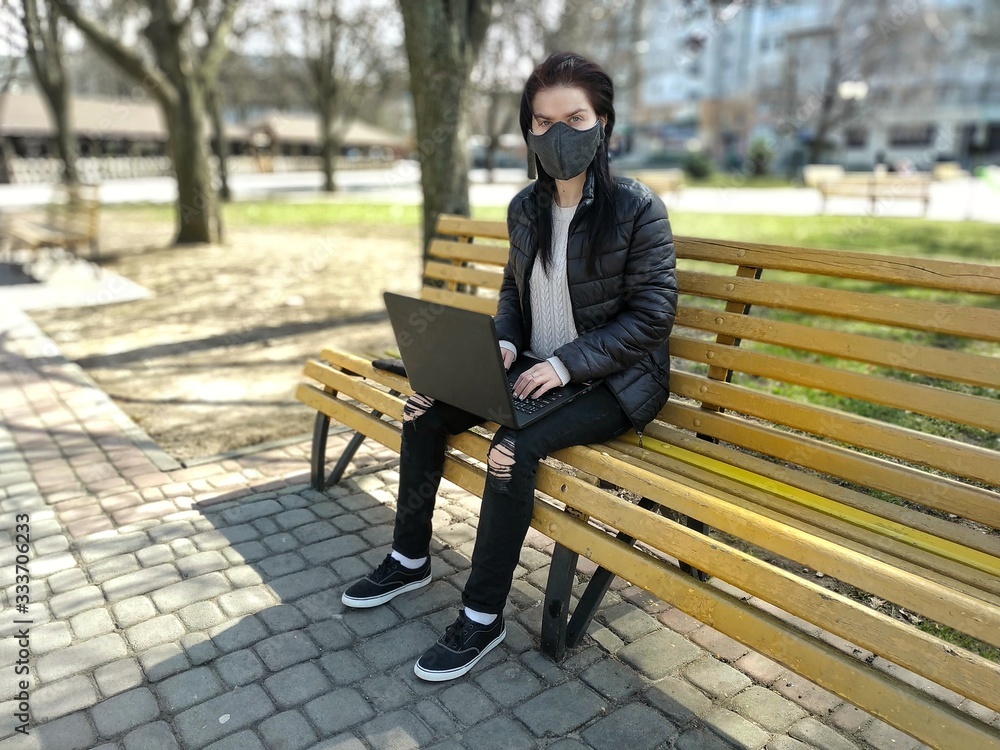 The masked girl is sitting on a bench in the park with a laptop and looking for work remotely during quarantine. Self-isolation and work remotely - problems with work, job search