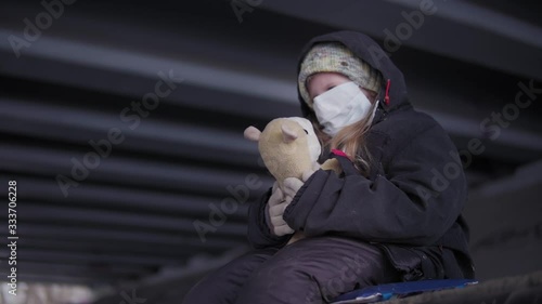 Little street kid with protective mask in black clothes and holey hat hugs toy under road bridge in the afternoon.