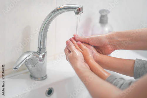 Woman washes the hands of a child with liquid soap over a white sink in in the bathroom. Closeup of a woman washing the hands of a child carefully with soap in the sink under running water.