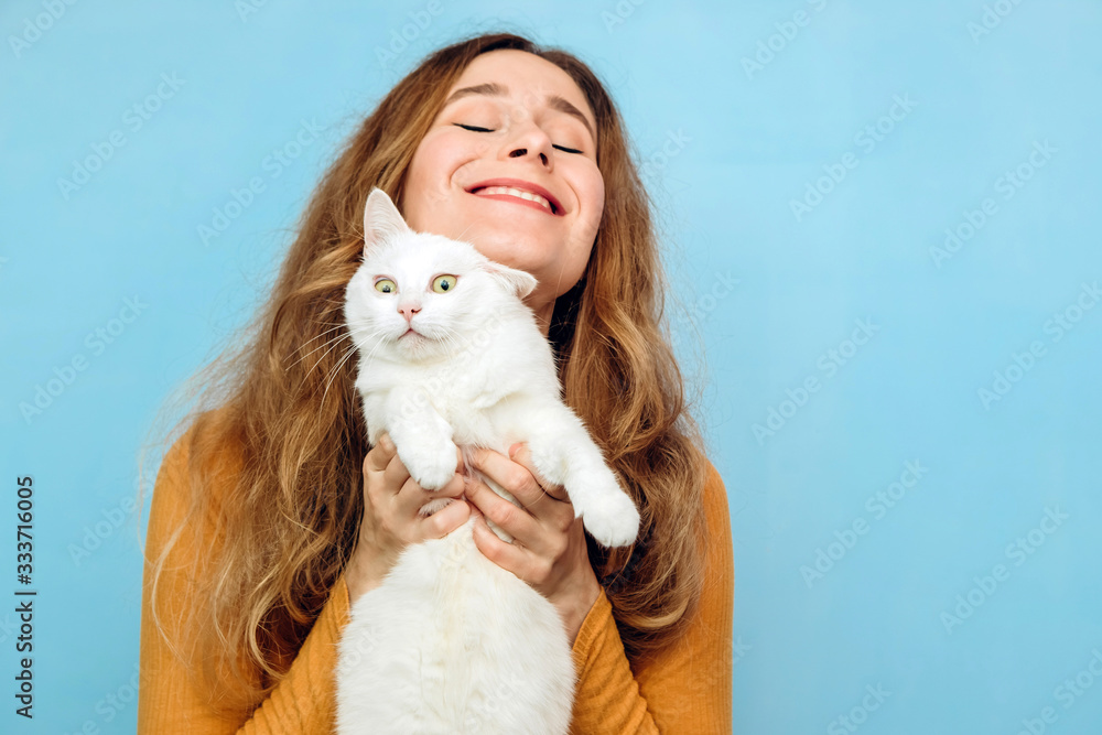 Obraz premium A young girl is holding a white cat in her arms. Portrait of a curly-haired blonde girl on a blue background. The concept of animal protection. Take the cat from the shelter.