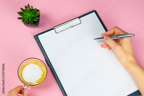 Tablet with white flattery next to which stands a green plant, hand holds a pen and writes on a sheet in the other hand a cup with a coffe photo