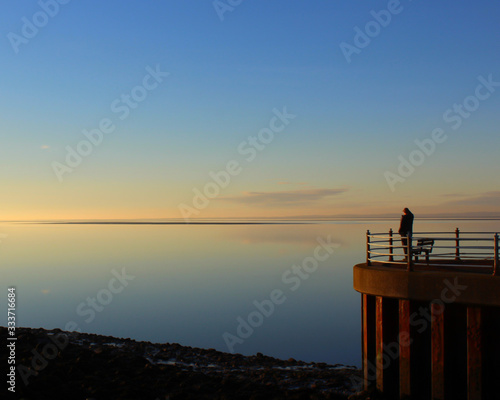 A lone figure stands looking out over the calm sea on a golden sunset at Morecambe in Lancashire