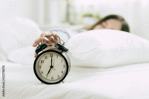 hand of young woman sleeping on bed pressing snooze button on black vintage alarm clock at seven o'clock morning in bed room at home, lifestyle, good morning, healthy sleep and joyful weekend concept