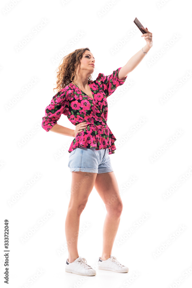 Fototapeta premium Side view of stylish fashionable young curly hair woman taking selfie with hand on hip. Full body isolated on white background. 