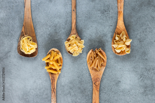 Different types of pasta lying in wooden spoons lying on grey table. Penne, tortellini, fuzilli, and farfalle. 