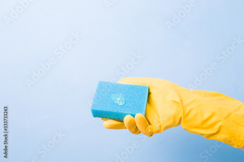 hand in a yellow rubber glove holds a blue sponge with a drop of green dishwashing gel, blue background copy space, cleaning concept, home care, spring cleaning