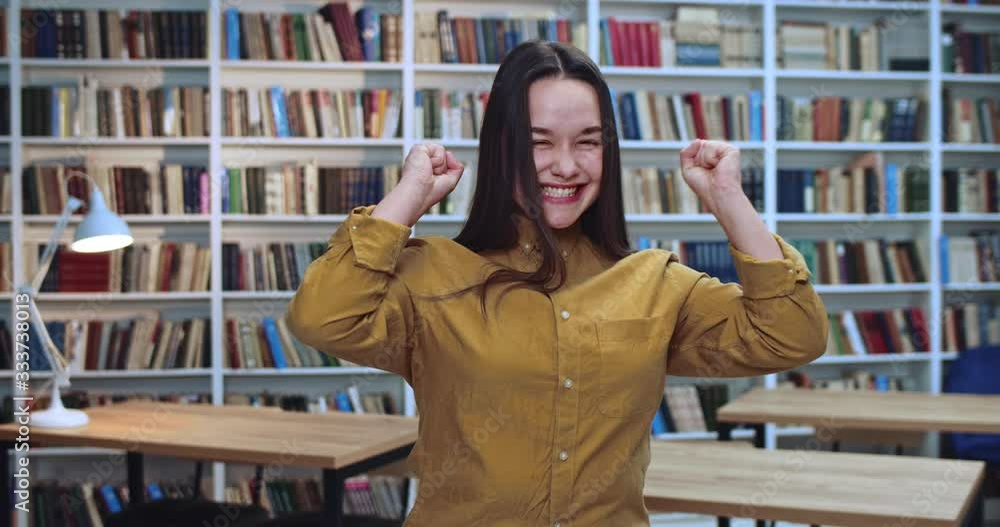 Portret of joyful brunette female student jumping and dancing in library after getting the best grade on exam. Portret of joyful brunette female student jumping and dancing in library after getting the best grade on exam.
