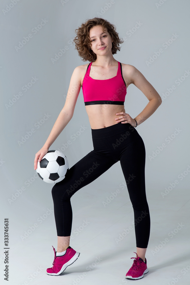 Full length, young athletic woman with a soccer ball in her hands, standing against a gray background and looking at the camera.