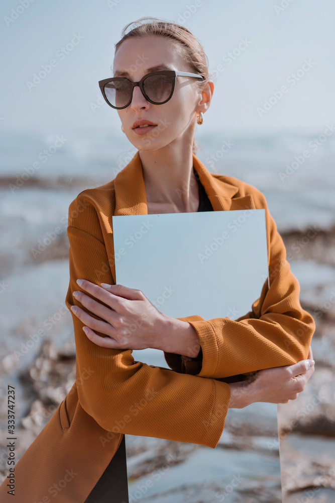 portrait Stylish girl in sun glasses on the sea beach holding mirror with reflection of blue sky. New vision of fashion projects concept. Dreams and illusions about travel