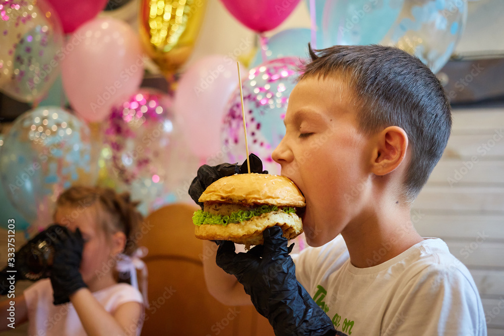 Naklejka premium The handsome little boy eating burger in black rubber gloves.