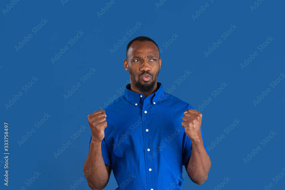 Fototapeta premium Celebrating win. Monochrome portrait of young african-american man isolated on blue studio background. Beautiful male model. Human emotions, facial expression, sales, ad concept. Youth culture.