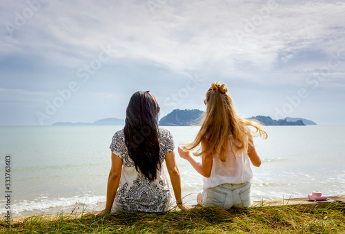 back of two female friends sitting on the beach enjoying sea view 