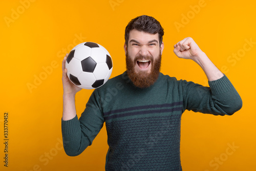 Excited bearded man holding a ball in his hand while screamin and making winner gesture on yellow background. photo