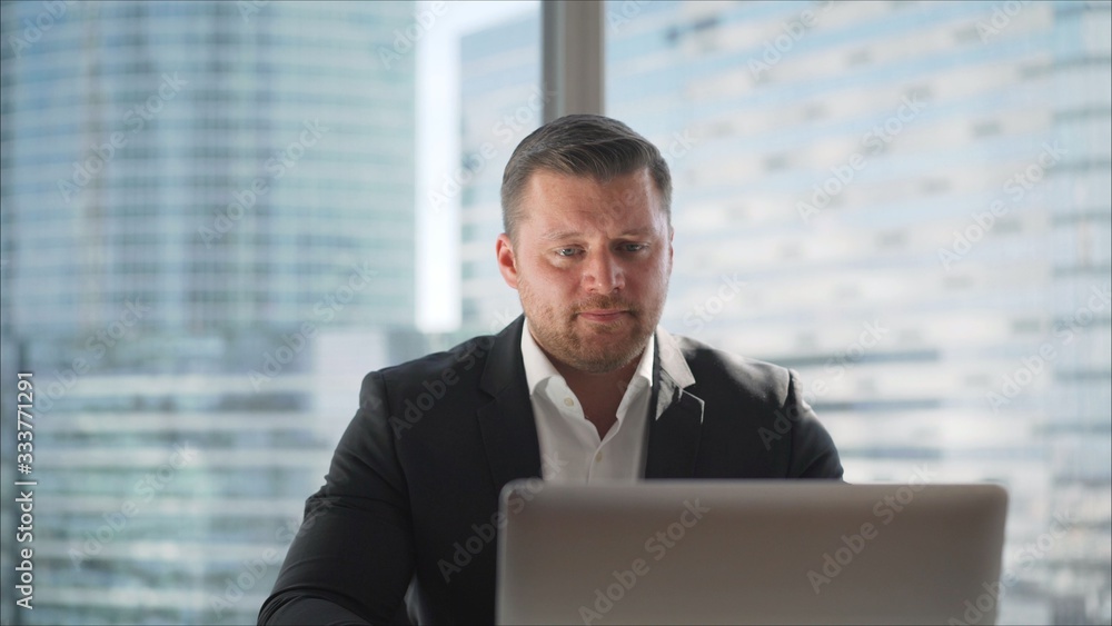 Young businessman in his office on the background of a skyscraper. Thoughtful young businessman in an elegant suit sitting on the background of a window in his office with skyscrapers 