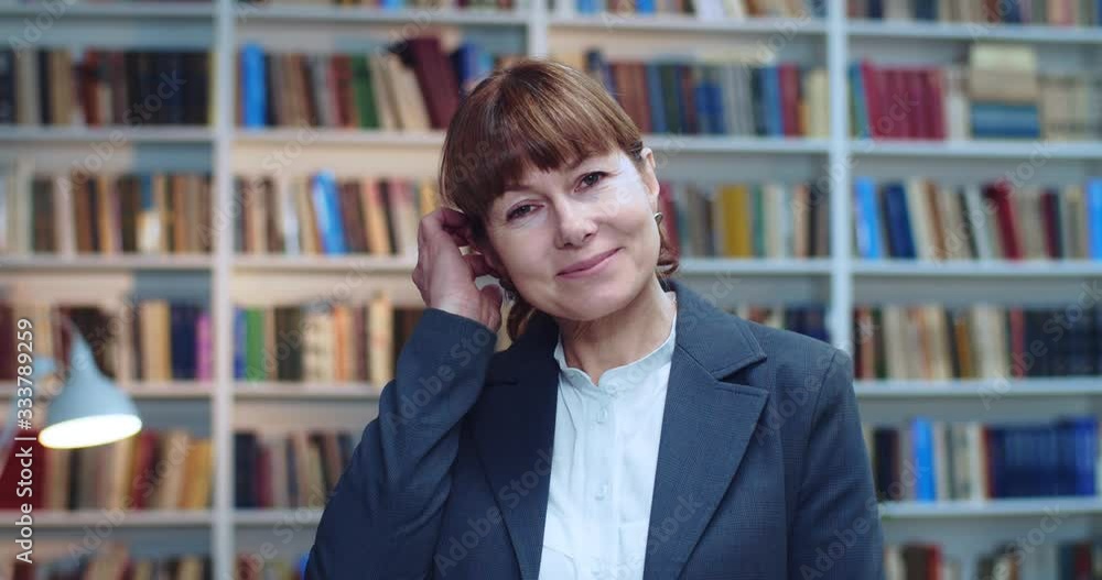 Close-up portrait of happy middle aged female professor standing in library next to book rack looking at camera and smiling while touching her hair. Close-up portrait of happy middle aged female professor standing in library next to book rack looking at camera and smiling while touching her hair.
