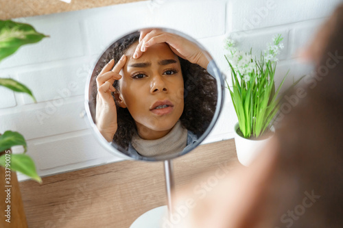 African teen girl feel worried about facial skin problem look in mirror. Young woman touch face squish pop zit pimple on forehead frustrated by blackheads. Black skin care problem, self care concept.