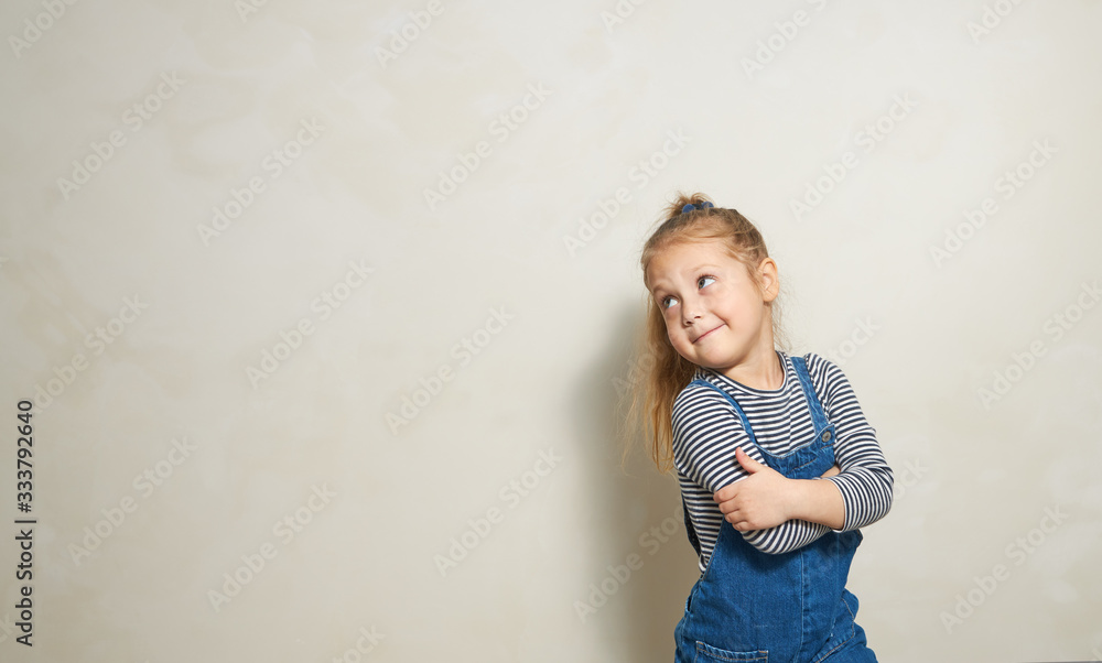 Funny little girl child stands near a white wall and looks with interest to the side. Space for text Funny little girl child stands near a white wall and looks with interest to the side. Space for text