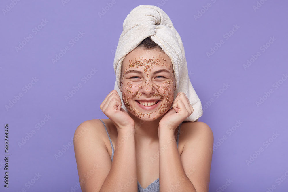 Studio shot of smiling happy female, model has rejuvenation procedure, cleans face with coffee peeling mask after taking shower, looks younger, posing isolated over lilac background. Studio shot of smiling happy female, model has rejuvenation procedure, cleans face with coffee peeling mask after taking shower, looks younger, posing isolated over lilac background.