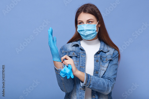 Image of darkhaired Caucasian woman with long hair, wearing medical face mask and putting on disposable gloves, looks at distance, standing isolated over blue background. Coronavirus, covid 19 concept photo