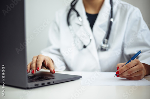 Closeup of a woman doctor wearing white medical gown and blue gloves  stethoscope. Female nurse sits at the desk with laptop and papers diagnosis. Coronavirus  covid-19  sickness prevention concept.