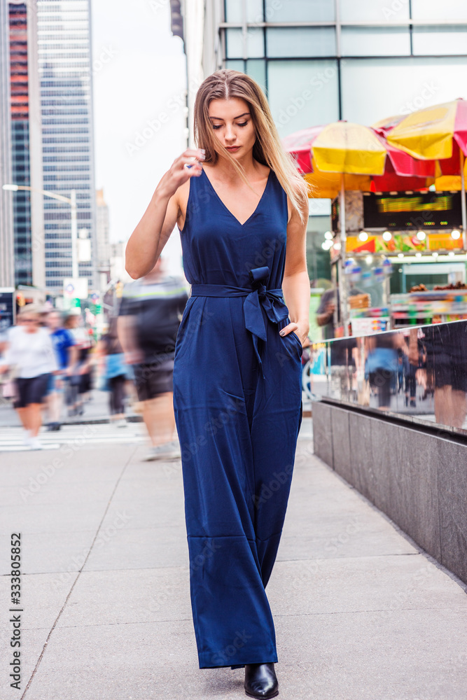 Fototapeta premium Young Woman Street Fashion in New York City. Young Eastern European Woman wearing blue sleeveless, v neck jumpsuit, black leather shoes, walking on busy street in middletown of Manhattan, relaxing..