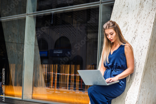 Young Eastern European American Woman working in New York, wearing blue sleeveless jumpsuit, standing against column outside office building, bending leg, looking down, working on laptop computer.