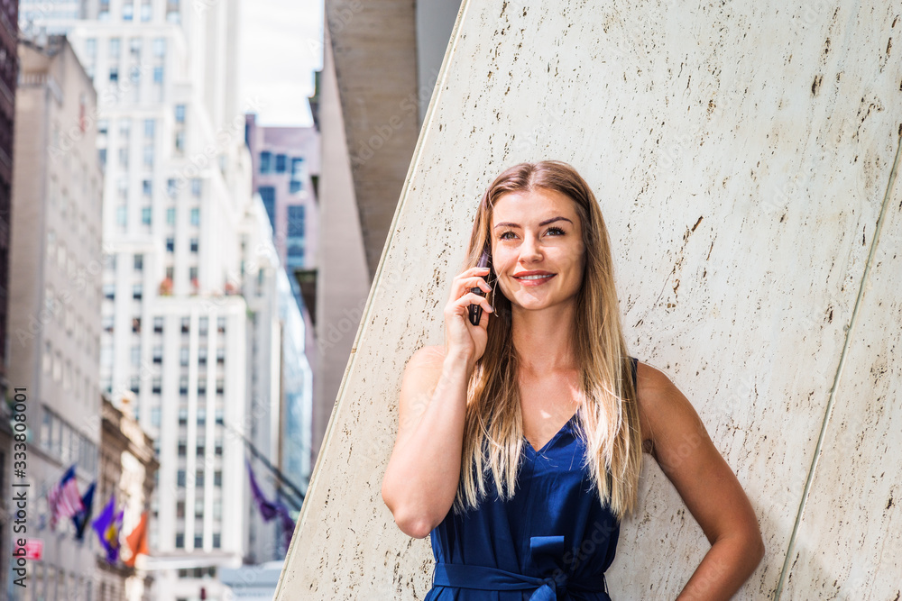 Young Eastern European American Woman with long brown hair, wearing blue sleeveless, v neck jumpsuit, standing against column outside office in New York City, talking on cell phone, smiling..