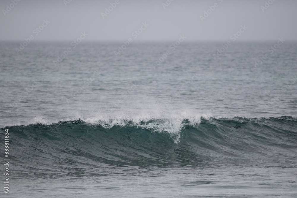 The start to a long wave with a spray coming over the top of the curl. It's a grey gloomy day. The sky and water are pale grey in color. The water is in both the foreground and background.