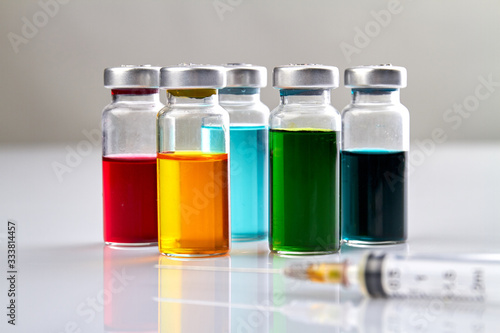 Glass vials with colored medicine liquid and syringe. White reflective surface.