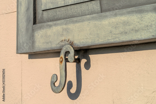 Close up of an antique shutter latch holding a green wooden shutter open against a beige building. 