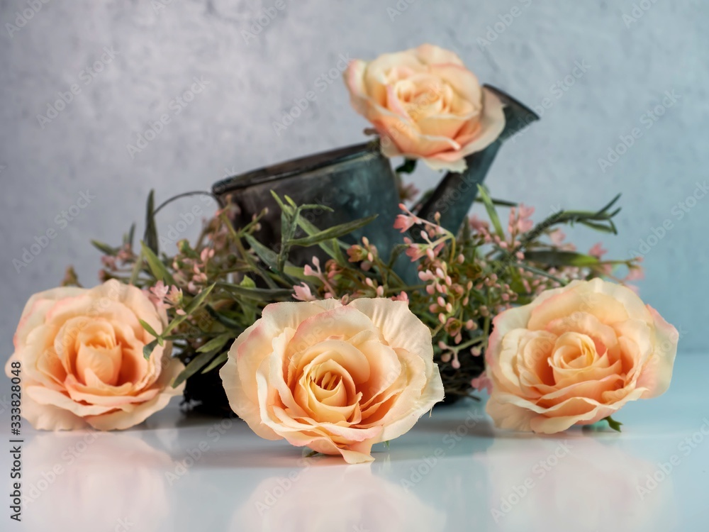 Obraz premium 4 peach roses and a floral pink and green wreath with a rustic metal watering can with a white counter and a plaster wall. Simple, soft, country home decor.