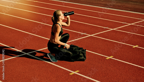 Young woman athlete in sportwear getting ready for workout while opening sports bag and drink water on the track of stadium at bright sunny day
