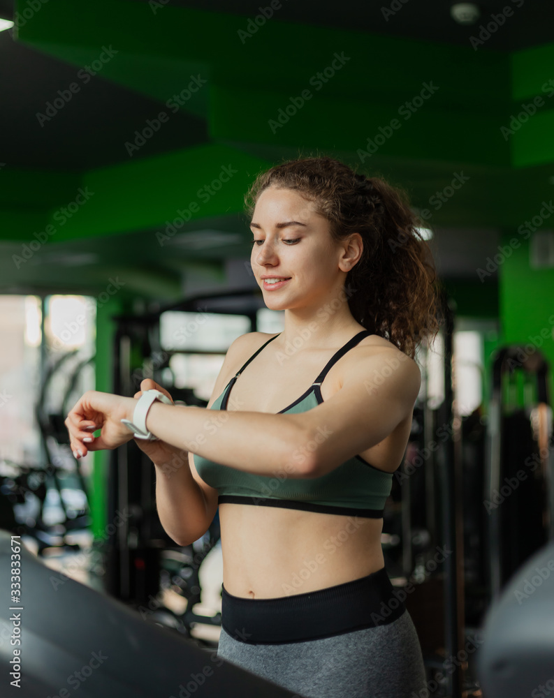 Fit woman in sportswear runs on a treadmill and looks at her watch in the gym. The concept of a healthy lifestyle, warming up, fitness, weight loss. Fit woman in sportswear runs on a treadmill and looks at her watch in the gym. The concept of a healthy lifestyle, warming up, fitness, weight loss.